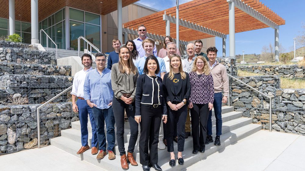 Fifteen people stand on steps outside a building and smile for the camera.