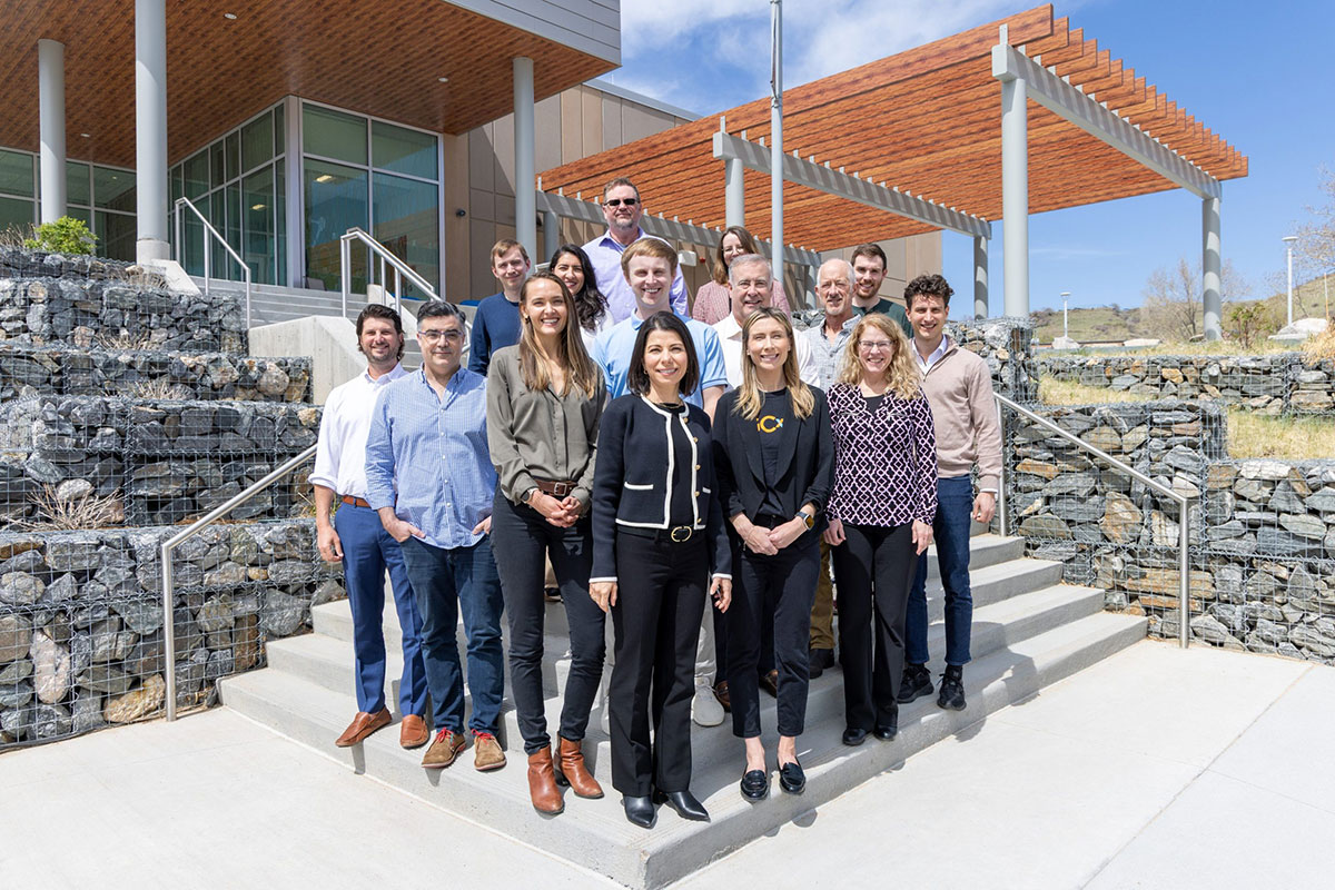Fifteen people stand on steps outside a building and smile for the camera.