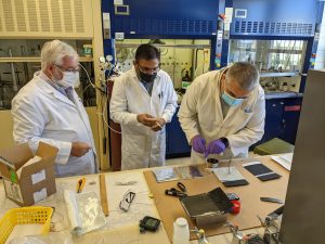 Two people in lab coats and masks watch as a third pipes something into a container placed on a table.